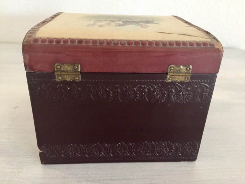 May include: A small wooden box with a burgundy lid and a floral design on the top. The box has brass hinges and a brown embossed design on the bottom.