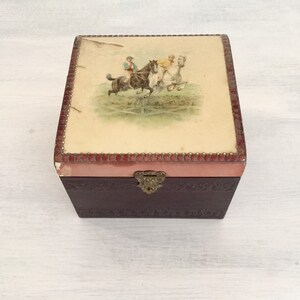 May include: A vintage wooden box with a hinged lid. The lid is decorated with a colourful illustration of three people riding horses. The box has a brown leather exterior and a pink interior.