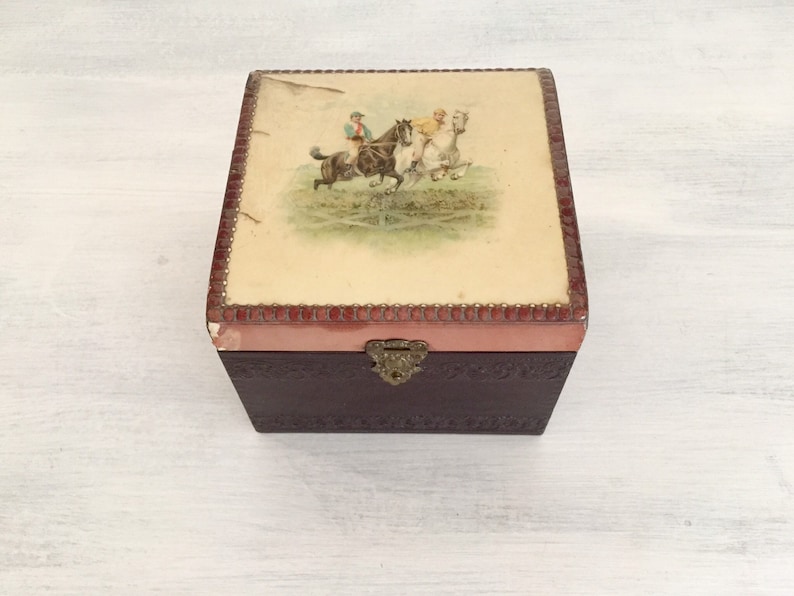 May include: A vintage wooden box with a hinged lid. The lid is decorated with a colourful illustration of three people riding horses. The box has a brown leather exterior and a pink interior.