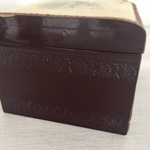 May include: A dark brown wooden box with a decorative embossed border and a cream-coloured top with a faded floral design.