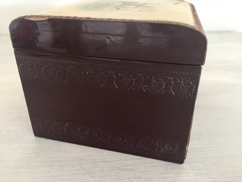 May include: A dark brown wooden box with a decorative embossed border and a cream-coloured top with a faded floral design.