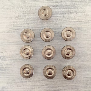 May include: Ten silver-toned round cabinet knobs with a raised center ring. The knobs are arranged in a pyramid shape on a white wooden surface.