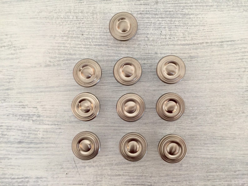 May include: Ten silver-toned round cabinet knobs with a raised center ring. The knobs are arranged in a pyramid shape on a white wooden surface.