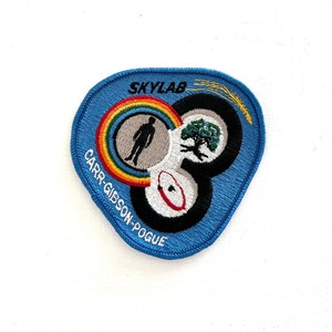 May include: A blue embroidered patch with the text "SKYLAB" and "CARR-GIBSON-POGUE" surrounding a design of three circles. The circles contain a rainbow, a stick figure, a tree, and a red ring.