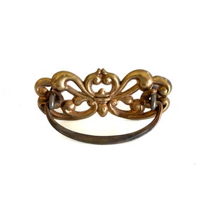 May include: A gold-toned ornate drawer pull with a decorative, openwork design and a curved metal handle.