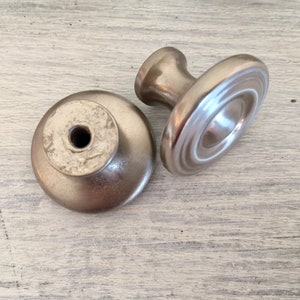 May include: Two silver cabinet knobs with a round base and a raised center. One knob has a smooth surface and the other has a textured surface.