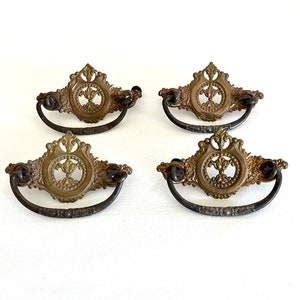 May include: Four antique brass drawer pulls with ornate floral designs and a ring handle. The pulls have a dark brown finish.