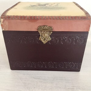 May include: A small wooden box with a hinged lid. The lid is decorated with a floral design and a pink border. The box has a brass latch and a brown finish.