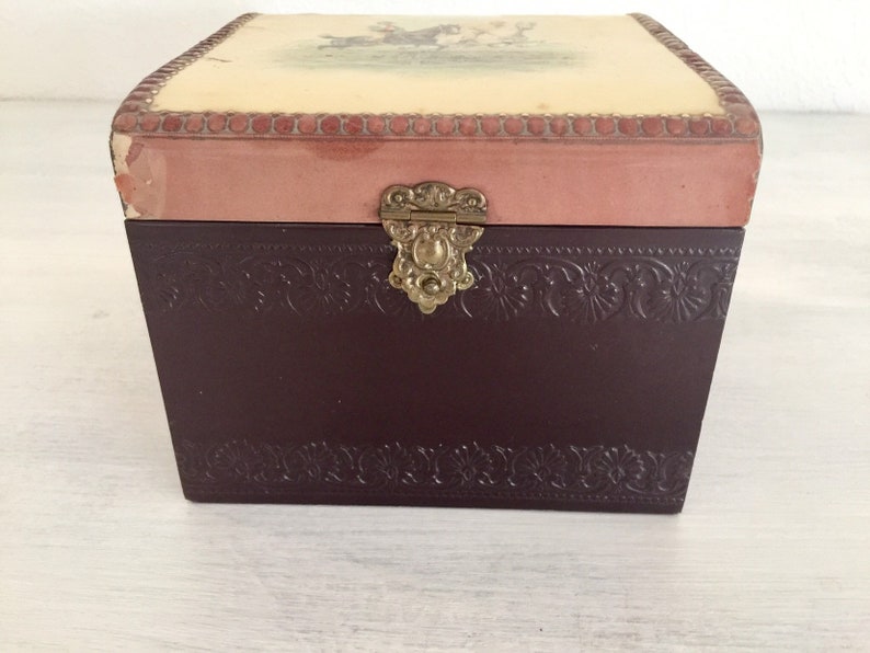 May include: A small wooden box with a hinged lid. The lid is decorated with a floral design and a pink border. The box has a brass latch and a brown finish.