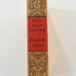 May include: A vintage book cover with a red spine. The title is "Hillbilly Doctor" by Elizabeth Seifert. The cover features a decorative floral design.