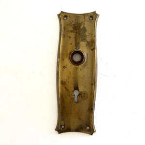 May include: Antique brass door plate with a keyhole and a circular opening. The rectangular plate has decorative edges and four screw holes. The brass has a weathered appearance, showing signs of age and use.