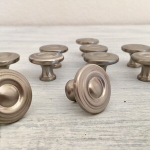May include: A set of ten silver-toned cabinet knobs with a circular design. The knobs have a raised center and a series of concentric circles around the edge.