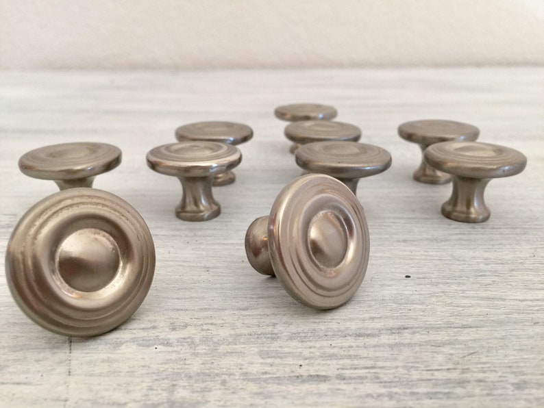 May include: A set of ten silver-toned cabinet knobs with a circular design. The knobs have a raised center and a series of concentric circles around the edge.