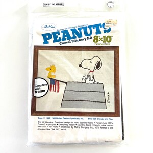May include: A Peanuts crewel embroidery kit featuring Snoopy and Woodstock standing on a doghouse with an American flag. The kit includes a pre-printed design on 100% polyester fabric, Persian type 100% Creslan acrylic yarn, an embroidery needle, a mounting board, and easy-to-follow instructions. The finished size is 8x10 inches and comes with a frame.