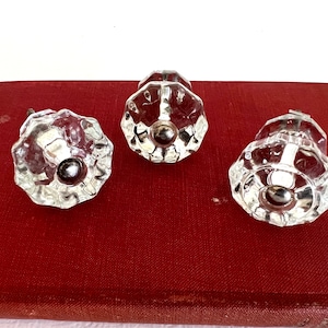 May include: Three clear glass cabinet knobs with a faceted design and a silver-colored center. The knobs are arranged on a red surface. These decorative knobs are suitable for furniture restoration or adding a vintage touch to cabinets.