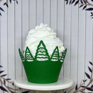May include: A green cupcake wrapper with a Christmas tree design holds a swirl of white frosting. The cupcake sits on a white pedestal, with a background of white vertical planks and decorative branches.