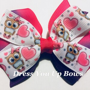 May include: A layered hair bow featuring a cute owl and heart pattern on white ribbon. The bow has a gradient of pink and purple, with a red center. The text "Dress You Up Bows" is printed on the bow.