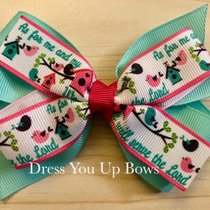 May include: A turquoise and pink hair bow with a decorative design. The bow features a white ribbon with a bird and birdhouse pattern and the text "As for me and my house, we will serve the Lord". The bow is from Dress You Up Bows.