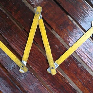 Pode incluir: Uma fita métrica dobrável amarela, com marcações de medição pretas, está disposta sobre uma superfície de madeira escura. A fita métrica está parcialmente aberta, exibindo números de 20 a 70 polegadas (aprox. 51 a 178 cm). A madeira tem um acabamento brilhante.