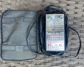 May include: A vintage camera flash unit with a silver reflector and the word "FILM" in red. The flash is black and has a black cord. A gray canvas case with a zipper is next to the flash.