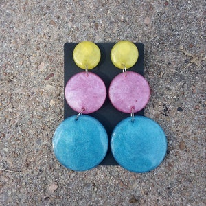 May include: A pair of handmade earrings with a three-tiered design. The top circles are yellow, the middle ones are pink, and the bottom ones are blue. The earrings are displayed on a black surface against a textured gray background.