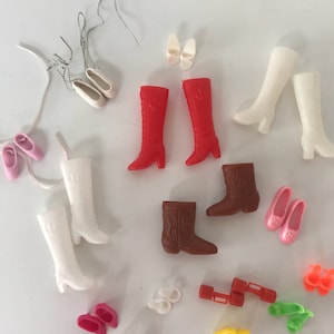 May include: A collection of vintage doll shoes and boots in various colors, including red, white, brown, and pink. The shoes are made of plastic and have different styles, such as cowboy boots, high heels, and flats.