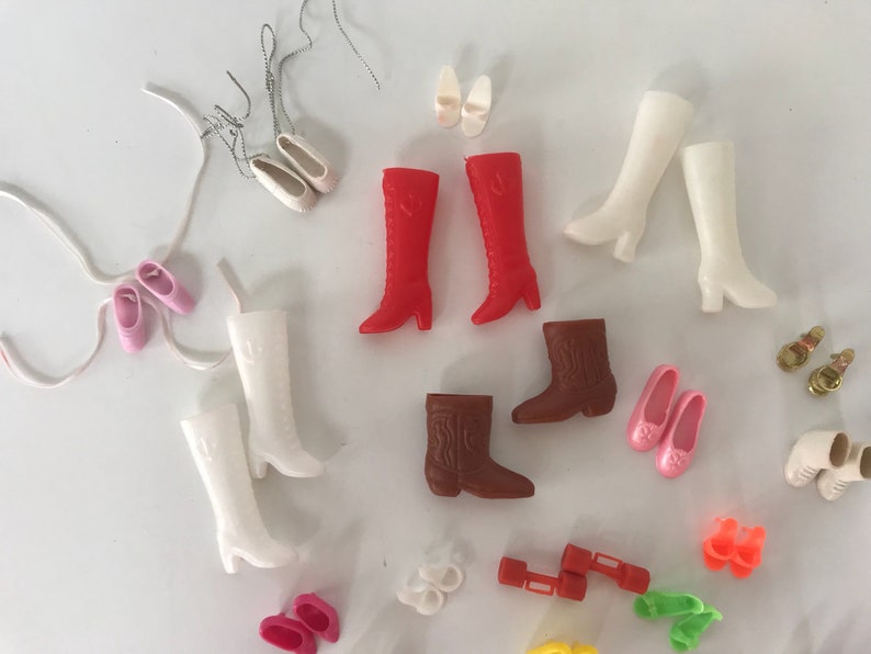 May include: A collection of vintage doll shoes and boots in various colors, including red, white, brown, and pink. The shoes are made of plastic and have different styles, such as cowboy boots, high heels, and flats.