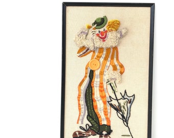 May include: A framed embroidered artwork of a clown with a striped orange and white costume, a green hat, and a red nose. The clown is holding an umbrella and is standing on a beige background.