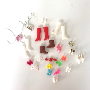May include: A collection of various doll shoes and boots in different colors and styles, including white, red, brown, and pink. The shoes are arranged on a white surface.