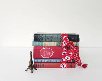 May include: A stack of vintage books tied with a red ribbon featuring white daisy patterns. The books have various colors and titles, including "SPY STORIES" and "THE WORLD'S BEST FAIRY TALES." A small Eiffel Tower figurine sits nearby.