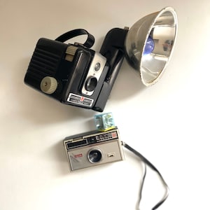May include: Two vintage cameras, a black and silver Brownie Hawkeye camera with a flash attachment and a silver Kodak Instamatic 104 camera with a black strap.