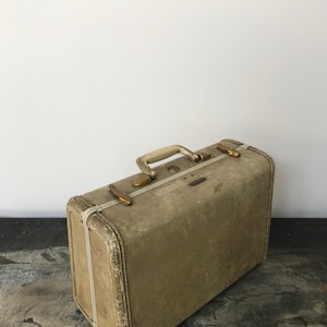 May include: A vintage beige suitcase with a white trim and brass hardware. The suitcase has a worn, distressed look.
