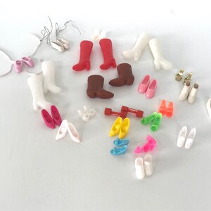 May include: A collection of various doll shoes and boots in different colors, including white, pink, red, brown, yellow, blue, and green. The shoes are made of plastic and are in various styles, including flats, heels, boots, and sandals.