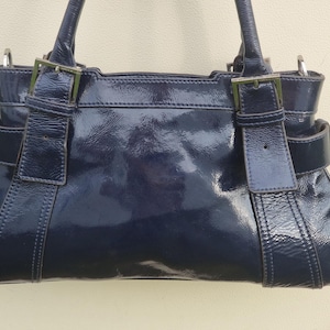 May include: A dark blue patent leather handbag with two top handles and silver hardware. The bag has a flap closure with a buckle and a structured shape.