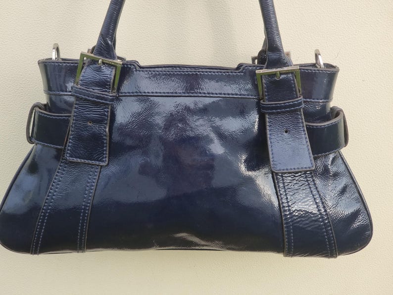 May include: A dark blue patent leather handbag with two top handles and silver hardware. The bag has a flap closure with a buckle and a structured shape.