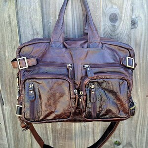 May include: A brown leather tote bag with multiple zippered pockets and adjustable straps. The bag has a glossy finish and features gold-colored buckles and zippers. The bag has two top handles and a shoulder strap.