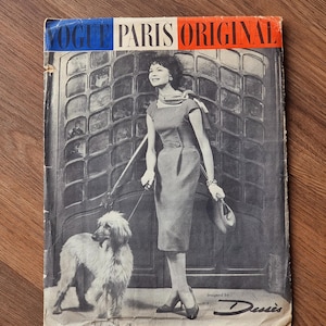 May include: A vintage Vogue Paris Original sewing pattern envelope featuring a black and white photograph of a person in a fitted dress holding a leash attached to a long-haired dog. The text reads VOGUE PARIS ORIGINAL and designed by Dessès.