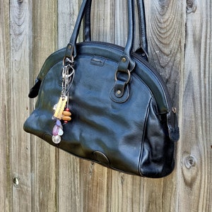 Könnte beinhalten: Schwarze Lederhandtasche mit rundem Oberteil und Doppelgriffen. Ein dekorativer Schlüsselanhänger mit Perlen und Holzakzenten hängt am Griff. Die Tasche hat einen Reißverschluss und ein kleines Logo.
