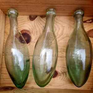 May include: Three vintage, clear glass bottles with a green tinge, displayed in a wooden crate. The bottles have a long, slender form with rounded bases and narrow necks. The glass shows imperfections and bubbles.