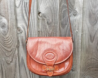 May include: A brown leather shoulder bag with a flap closure and a metal buckle. The bag has a small, round, embossed logo on the front.