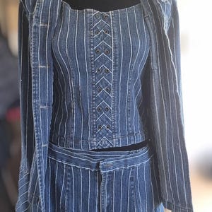 Könnte beinhalten: Ein dreiteiliges Jeans-Outfit mit vertikalen weißen Streifen. Das Set beinhaltet ein kurzes Korsett-Top mit Schnürdetail, eine passende Jacke und eine hoch taillierte Hose. Das Outfit wird auf einer schwarzen Schaufensterpuppe präsentiert.