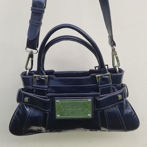May include: A dark blue leather handbag with silver hardware and a top handle. The bag has a flap closure and a small, rectangular metal plate with the brand name.