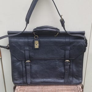 Könnte beinhalten: Schwarze Lederaktentasche mit Tragegriff und Schulterriemen. Die Tasche hat zwei vordere Schnallen und ein kleines Etikett mit der Aufschrift "HIDESIGN". Die Tasche steht auf einem Korbstuhl.