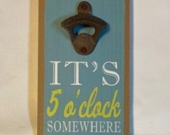 May include: A wooden wall-mounted bottle opener with a blue background and the text "It's 5 o'clock somewhere" in yellow and white. A yellow cocktail glass with a green umbrella is pictured below the text.