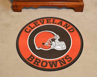 May include: Round rug featuring the Cleveland Browns logo. The rug has a black center with an orange football helmet and the words "Cleveland Browns" in white and orange. The rug is on a light brown carpet.