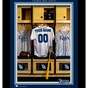 May include: A framed print of a baseball locker room with a white jersey with the text "YOUR NAME" and the number "00" hanging on a hook. The jersey is flanked by two blue and white jerseys with the word "RAYS" on the front. The locker room has wooden lockers with the letters "TB" on the front. The print has a blue border with the text "TAMPA BAY RAYS™" at the top and the text "FAN CREATIONS/MLB" at the bottom.