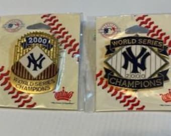 May include: Two gold and blue New York Yankees World Series Championship pins from 2000. The pins are on a red and white baseball-themed background.