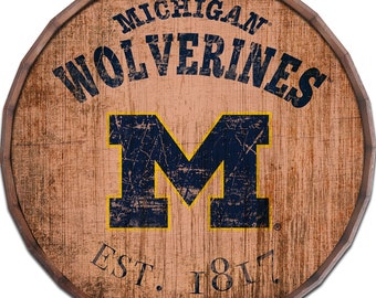 May include: A round wooden sign with a distressed woodgrain finish. The sign features the University of Michigan Wolverines logo, a blue "M" with a yellow outline, and the text "Michigan Wolverines Est. 1817".
