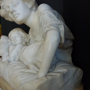 May include: A white marble sculpture of a woman holding a sleeping child. The woman is wearing a flowing robe and the child is lying on a pillow. The sculpture is on a pedestal.