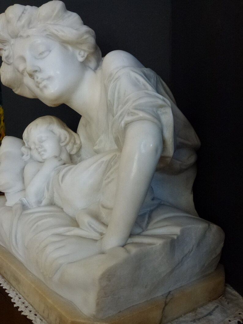 May include: A white marble sculpture of a woman holding a sleeping child. The woman is wearing a flowing robe and the child is lying on a pillow. The sculpture is on a pedestal.